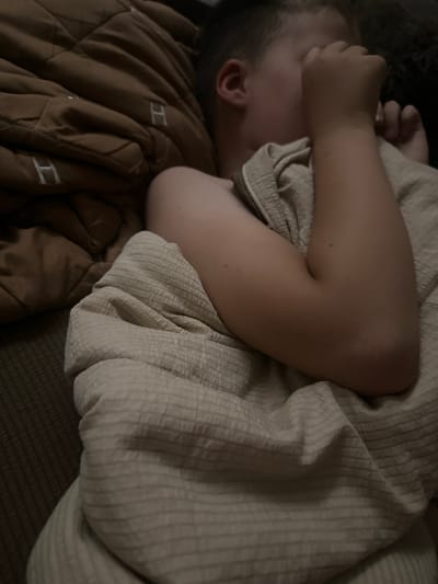 A person is sleeping in bed, curled up on their side and covering their face with their arm. The person is under a light-colored, textured blanket. The bed is covered with a brown, quilted comforter with white embroidered "H" symbols. The scene is dimly lit, suggesting it might be nighttime or early morning. The overall impression is one of peaceful rest. No weather cues are visible. The location context of Peremyshliany, Ukraine is not discernible from the image itself.