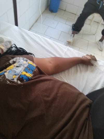 The scene is set in what appears to be an institutional room, likely a medical facility, in Mape, Nigeria, characterized by white tiled walls and a white tiled floor with visible grime in the grout. In the foreground, a person lies supine on a bed covered with white sheets, viewed from above. The individual wears a brown t-shirt featuring a multi-colored graphic (yellow, blue, and grey) on the left chest. Their dark, braided hair rests on a pillow. A light-colored bandage or plaster is affixed to the inner forearm/wrist area of their right arm, which extends down the bed. Dark clothing is visible to the right of the person's torso. In the background, a second individual stands, only their lower body visible. They wear black pants, with a white athletic shoe on their right foot and a light-colored high-top shoe or boot on their left foot. A partial white chair or stool is visible behind their legs. Against the far wall, a blue bucket rests on the floor, and a white cable or cord runs vertically down the wall. A dark vertical structural element, possibly a beam or pipe, is present on the left side of the frame.