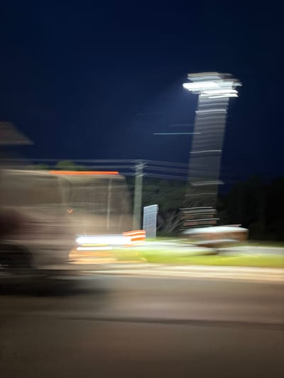 The image depicts a blurry, motion-induced scene at night, likely captured from a moving vehicle. A tall, illuminated structure, possibly a light pole or tower, dominates the right side of the frame, its light source appearing as a horizontal streak of white. To the left, a vehicle is visible, its lights creating streaks of orange and white. The background consists of dark, indistinct shapes suggesting trees and a deep blue night sky. The ground is a blurred expanse of asphalt. No specific landmarks of Venice, United States are discernible due to the motion blur and nighttime setting.