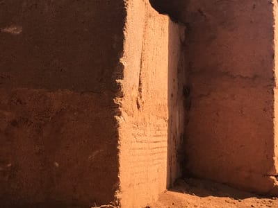 A rough, earthy structure features a square recess or niche in the left. The niche's interior and surrounding walls are textured, with varying tones of brown and tan. Light casts a strong shadow on one side, highlighting the uneven surface. Faint horizontal lines are visible on the wall beside the niche. The overall impression is of an aged, hand-built structure.