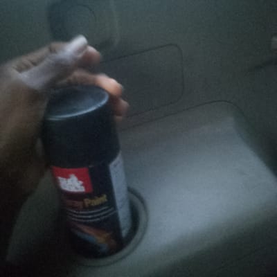 A hand holds a can of black spray paint above a sink or basin. The scene is dimly lit and the background is a light gray or beige color. The location (Isunba, Nigeria) is not visually discernible in the image.