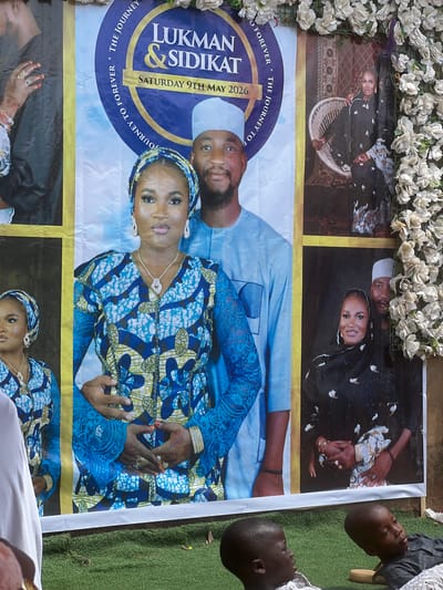 A rectangular poster, potentially laid on the ground outdoors, appears to commemorate a wedding or similar event. The poster features several images of a couple, along with the names "Lukman & Sidikat" and the date "Saturday 9th May 2026." The overall color scheme of the poster is predominantly blue and yellow. The poster appears to be bordered by a garland of artificial white flowers along the top. Portions of people are visible along the periphery of the image, suggesting the presence of others at the scene. The poster is likely located outdoors with grass visible nearby. The city and country of Rigasa, Nigeria is stated in the prompt, but not identifiable in this image.