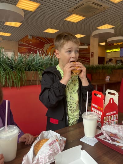 An interior shot depicts a young boy eating a burger at a fast-food restaurant in Krilovo, Russia. The boy, with light brown hair, wears a light green t-shirt adorned with game controller graphics under a black zip-up jacket. He is positioned slightly to the right of the frame, actively taking a bite from a sesame seed bun burger held in both hands, with his mouth open and eyes looking upward and left.

The boy is seated at a dark brown laminate tabletop. To his left, a partial view of another individual's hand and a purple sleeve are visible, alongside a clear plastic cup with a white liquid (likely a milkshake) and a pink straw. Further left, a burger wrapped in white paper with "CHICKEN" branding rests on the table. To the boy's right, a similar milkshake in a plastic cup with a pink straw is present, along with a printed receipt. Two red and white cardboard boxes, branded "CHICKEN детям" (CHICKEN for children) and "НАЙДИ ПАРУ" (Find a Pair), stand on the table, which also holds a red plastic tray and other food wrappers.

The background features red booth seating and a decorative wooden planter containing artificial green and light green foliage, separating dining areas. A large red and orange geometric mural adorns a distant wall. In the midground, beyond the foliage, another dark table and chairs are visible, along with blurred figures of other patrons or staff. A prominent yellow sign with Cyrillic text "ЗАКАЖИ ЗДЕСЬ" (Order Here) is visible in the upper right background. The ceiling is a grid-style drop ceiling featuring multiple square, yellow-orange illuminated light panels, several white circular pendant lights, and a square white air conditioning unit. The overall environment is well-lit and appears to be a modern, clean fast-food establishment.