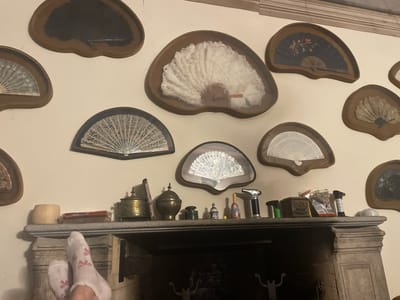 The image depicts a dimly lit indoor scene, likely a living room, focusing on a fireplace mantle adorned with various decorative fans and objects. The fans are displayed on the wall above the mantle, enclosed in brown, heart-shaped frames. They vary in size, material, and design, including a large white feather fan, a black fan with floral embroidery, and several lace or paper fans.
On the stone mantle, an assortment of items is arranged. These include what appear to be antique brass containers, a small green inkwell, a miniature lighthouse-shaped object, a matryoshka doll, a butane torch, and packets of cigarettes. A wooden cup-like object sits to the left of these items. In the foreground, a pair of feet wearing white socks with pink bows are visible, resting on the edge of the mantle. The fireplace itself is dark and appears to be unlit. The wall is painted a neutral color, and the room is decorated with a molding near the ceiling. The overall impression is of a collected display of antique and personal items. The scene does not provide any specific visual cues to definitively place it in Borgosesia, Italy.