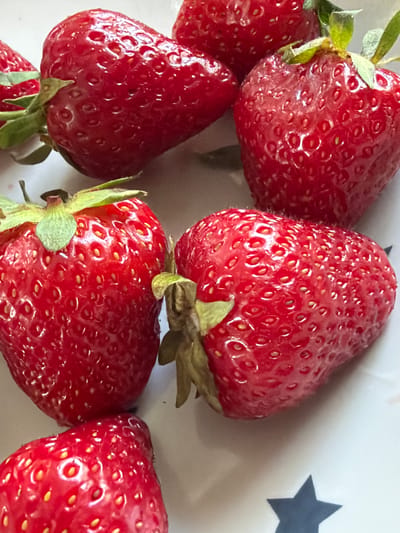 The image shows a close-up, overhead view of several ripe strawberries arranged on a white plate with a dark blue star pattern. The strawberries are a deep red color and appear fresh, with visible seeds. The lighting is bright and highlights the texture of the fruit. No people or actions are depicted. The specific location in Кирпичного завода № 1, Russia is not determinable from the visual information provided.