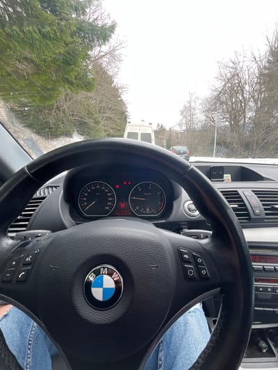 The image captures a first-person perspective from inside a BMW vehicle, depicting a winter road scene in Пампорово, Bulgaria. The driver's legs, clad in blue denim jeans, are visible at the bottom of the frame beneath a black, multi-function BMW steering wheel featuring the brand's emblem. The dashboard's instrument cluster displays a speedometer reading 0 km/h and a tachometer showing 0 RPM. Several red warning lights are illuminated, including battery and seatbelt indicators, and the odometer shows 191531 km. To the right on the dashboard, a black phone holder and a small framed picture are present.
Beyond the windshield, the snowy roadside is visible, leading to a paved road ahead. A white cargo van with orange roof lights is positioned directly in front of the BMW, appearing stationary or moving slowly. A dark-colored passenger car is partially visible behind the van. The environment is characterized by bare deciduous trees and green coniferous trees lining both sides of the road, indicative of a mountainous, wintry landscape. The sky is overcast and bright.