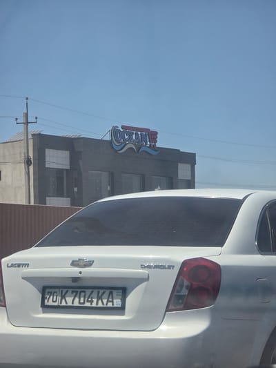 A white Chevrolet Lacetti sedan is parked in front of a modern, grey building with large windows. The building has a neon sign that reads "OCEAN". The license plate on the car is "70K704KA" and the country code "UZ" is visible. The scene appears to be in a city, possibly Yukari-Gazbiran, Uzbekistan, given the license plate format. The sky is clear and blue, suggesting a sunny day.