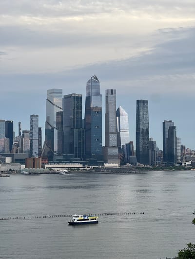 This image, taken from Weehawken, United States of America, captures a wide view of the New York City skyline across a body of water. The sky is overcast with soft, muted clouds. In the foreground, a ferry with blue and yellow markings is moving across the water. The water itself is calm and reflects the gray sky. Along the shoreline of the city, numerous skyscrapers of varying heights and architectural styles dominate the scene. Some buildings are clad in glass, reflecting the cloudy sky, while others have darker facades. A long, low-rise building complex with multiple white roofs is visible closer to the water's edge, with several smaller boats docked along it. In the distance, the cityscape extends further, with more buildings visible, some appearing as silhouetted shapes against the horizon. The overall impression is of a large, bustling urban environment on a cloudy day.