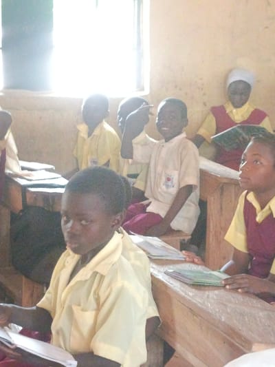 The image captures an indoor classroom setting within the city of Mada, Nigeria. Approximately seven students, appearing to be elementary school age, are visible. They wear light yellow short-sleeved uniform shirts; some attire also incorporates maroon or red elements such as trousers, vests, or head coverings. In the foreground left, a male student in a light yellow shirt holds an open notebook and looks directly towards the viewer. Behind him, another male student in a light yellow shirt is seated, smiling towards the viewer, with his right hand raised and index finger extended. To the foreground right, a female student in a yellow shirt and maroon top is partially visible, looking upwards, her hands resting on a stack of books on a wooden desk. In the background, other students in similar uniforms are seated at wooden desks and benches; one female student wears a white head covering and examines an open book. The classroom contains wooden desks and benches. Natural light enters from a large, bright window positioned on the left side of the frame. The walls are light-colored and exhibit signs of wear. Books and notebooks are present on the desks.