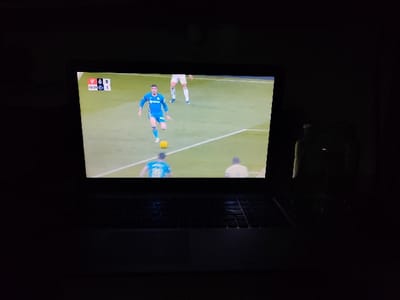 In an indoor setting in Rishtan, Uzbekistan, a silver-colored laptop is prominently displayed in a low-light environment. Its screen is the primary light source, illuminating a live soccer (football) match. The game is shown on a green field with white markings. A player in a blue jersey with white trim, dark hair, and light skin is the central figure, actively dribbling a yellow soccer ball towards the right side of the screen. Another player in a light blue jersey is visible in the foreground with their back to the viewer, while a player in a white jersey is partially seen in the background. An on-screen overlay displays game information: "00:21" (time elapsed), a score of "0" for the team on the left (indicated by a red emblem), and "1" for the team on the right (indicated by a black and white emblem). The laptop's keyboard and touchpad are visible in the foreground, with dimly lit keys. To the right of the laptop, a dark, cylindrical object, possibly a bottle or thermos, is dimly discernible, catching subtle reflections from the screen. The immediate surroundings are dark, suggesting nighttime or a room with minimal ambient light.
