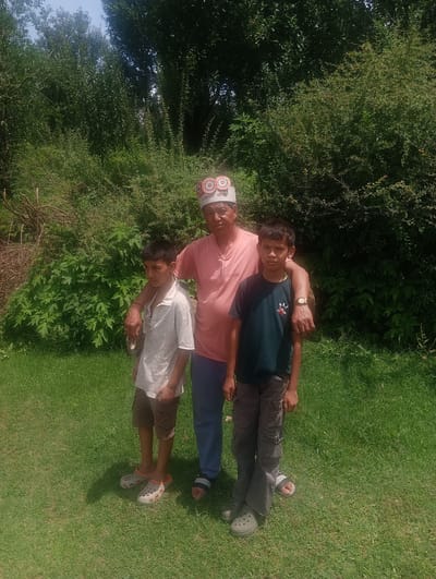 A man and two young boys stand on a grassy area in front of dense green foliage, suggesting an outdoor location in Khakhnal, India. The man, wearing a pink t-shirt and a decorative hat, has his arm around the shoulder of the boy to his left. This boy is wearing a light-colored, patterned shirt and shorts. The boy on the right is wearing a dark green t-shirt and gray cargo pants. Both boys are wearing casual footwear. The scene is illuminated by natural sunlight.