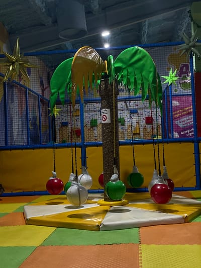This is a photograph taken inside an indoor children's playground in Kamyshlov, Russia. The scene is dominated by a faux palm tree structure in the center, from which multiple colorful, spherical punching bags hang by ropes. The punching bags are red, green, and white, and appear to be made of a durable material. The palm tree has green and yellow leaf-like decorations at its top. The playground floor is covered with interlocking foam mats in various colors including orange, yellow, and green. In the background, a blue safety net encloses a climbing area, and colorful graphics are visible on the wall, including what appears to be a large "A" in a stylized font. There is a sign on the palm tree trunk with a red circle and a diagonal line through it, indicating a prohibition, along with text in Russian. The lighting appears to be artificial, creating some shadows. No people are visible in the image.