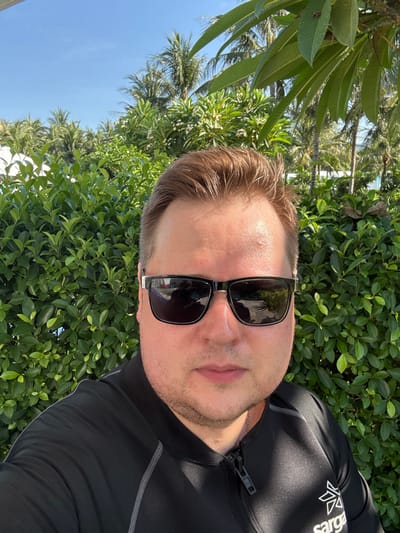A man is taking a selfie outdoors, with a backdrop of lush green bushes and palm trees under a clear blue sky. He is wearing a black rash guard with a zipper at the neck, and black sunglasses. The logo "sarga" is visible on the lower right side of his shirt. The environment suggests a tropical or subtropical location, possibly a resort or beach area in Cam Lâm, Vietnam. The lighting indicates it is daytime and sunny.