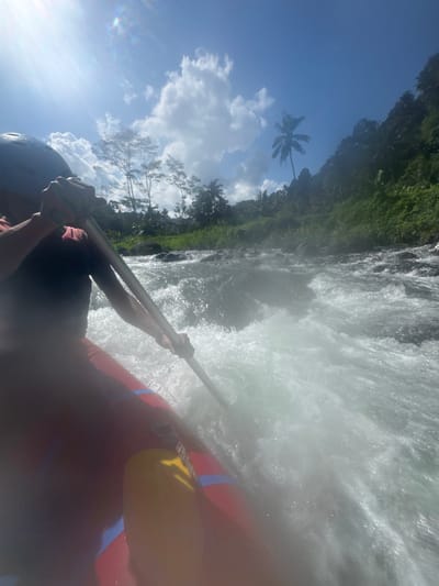 This is a first-person perspective, low-angle, medium shot of whitewater rafting. The image is vertically oriented. In the foreground, a person wearing a dark helmet and a dark shirt is holding a paddle. Part of a red and blue raft is visible. The raft is navigating through turbulent, foamy white water.
The background reveals a lush, green jungle-covered landscape with tall trees, including palm trees, against a bright blue sky dotted with white clouds. Sunlight glints off the water and creates a lens flare in the upper left portion of the image. The scene suggests a tropical location, consistent with Sidemen, Indonesia. The whitewater suggests an adventurous activity. The overall mood appears energetic and exciting due to the action of the rafting. The image quality is affected by water splashes, creating a slightly blurred and hazy effect. No text is visible in the image.
