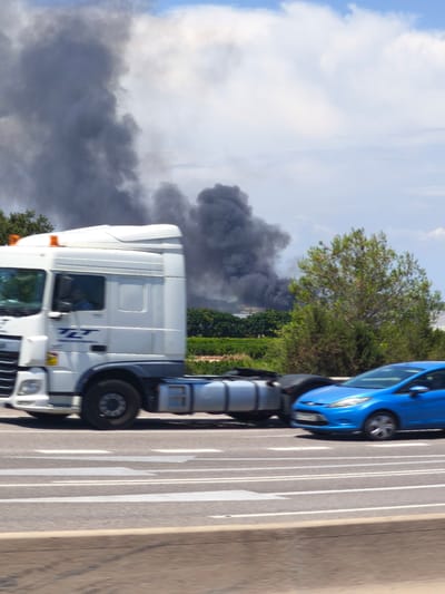 A white semi-truck is driving on a highway in Riba-roja de Túria, Spain. A blue car is driving in the lane next to the semi-truck. In the background, a large plume of black smoke is rising into the sky, indicating a fire. The sky is blue with some clouds. The environment appears to be rural or semi-rural, with trees and some vegetation visible. The image is taken from a low angle, looking up at the vehicles and the smoke. The overall impression is one of an ongoing emergency or accident.