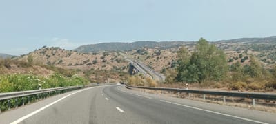 A wide-angle, high-angle shot depicts a two-lane asphalt road curving through a hilly, arid landscape under a clear, light blue sky. The road, marked with white dashed lines and a solid white line on the left edge, has visible tire tracks near the center. On the left side of the road, a metal guardrail runs along the edge, with a dense growth of green bushes and white flowers just beyond it. On the right side, a similar metal guardrail is present, with dry, yellow-brown vegetation and scattered trees behind it. In the mid-ground, a multi-lane highway with a prominent concrete overpass or viaduct is visible traversing the terrain, with several vehicles on it. The surrounding hills are covered in sparse, dry vegetation and dotted with scattered trees. The distant background shows more rolling hills with similar arid vegetation and a few scattered trees. The overall impression is a remote, dry, Mediterranean climate. The image is taken from a vehicle moving along the road, suggesting a travel or transportation context. The location is identified as El Garrobo, Spain.