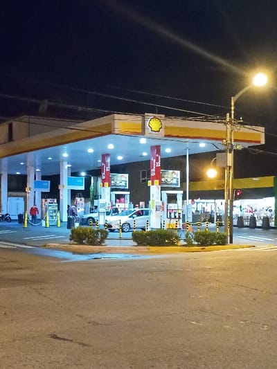 The image shows a Shell gas station at night, located at -34.453, -58.539 in the city of Victoria, Argentina. The gas station is brightly lit with overhead lights, illuminating a gray sedan at one of the pumps. There is a single employee in the foreground, partially obscured by a gray sedan. The employee appears to be wearing a blue shirt and jeans.  The gas station is surrounded by green bushes and yellow painted curbs, with a dark street in the foreground. The street has a crosswalk visible. There is a streetlight on the right side of the image, illuminating the scene. The image was taken from a street level perspective, with the gas station visible in the distance.  There is an additional streetlight visible on the right side of the image and the gas station canopy has a "Shell" logo in the middle. The gas station has several pumps, all displaying a bright yellow color. The sign on the pump shows the price per liter for different types of fuel.  There are no people visible in the pumps. The image is in focus, with the gas station and the surrounding environment well-lit and visible.  The image was taken at night, with the streetlights illuminating the scene. The image was taken in a horizontal orientation. There is a stop sign on the right side of the image. The image was taken at night, but the streetlights have illuminated the image well.