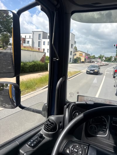 The image presents a first-person view from the interior of a vehicle, likely a truck or large commercial vehicle, looking out onto an urban or suburban street in Quickborn, Germany, during the daytime. The sky above is partially cloudy but bright.

Inside the vehicle, the black steering wheel is prominently positioned on the right side of the frame. The dashboard features a speedometer indicating speeds up to 120 km/h, with the needle hovering between 30 and 40 km/h. A digital display shows "10:57" and "33°C," alongside other text such as "Bağlı değ" (Turkish for "not connected") and "Telefon." Controls for lights and a steering wheel-mounted "OK" button with directional arrows are visible. On the door panel, there is an air vent and a control module for window and mirror adjustments. Large side mirrors, characteristic of a truck, reflect parts of the vehicle's yellow body and the surrounding environment, including buildings and trees.

Outside the vehicle, a paved road with white lane markings stretches forward. A dark-colored car, possibly a BMW, is ahead in the lane, with another vehicle visible further in the distance. A red traffic light is active in the distance, suggesting the vehicle may be stopped. Construction barriers, white and red, are also faintly visible further down the road. Along the roadside are various buildings: modern, multi-story structures with white and grey facades and prominent windows, one featuring a green roof, and an older, yellow building with a red roof displaying a circular "50" sign. Greenery, including trees, bushes, and grass, lines the street, with a manhole cover visible on a grassy strip. Reflections on the windshield include the word "Hamburo" and a "90" speed limit sign. The overall scene depicts a mundane moment of travel through a town.