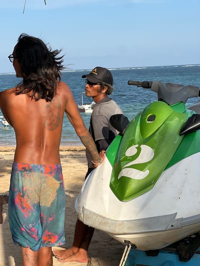 Two men stand on a sandy beach next to a white and green jet ski. The man on the left has long dark hair, is wearing patterned swim trunks, and has tattoos on his back and arm. He is facing away from the camera. The man on the right wears a black baseball cap and a dark t-shirt. He is looking to his left. The jet ski is parked on a blue trailer. The ocean is visible in the background with small waves breaking. The sky is clear and blue. The scene appears to be a public beach in Denpasar, Indonesia.