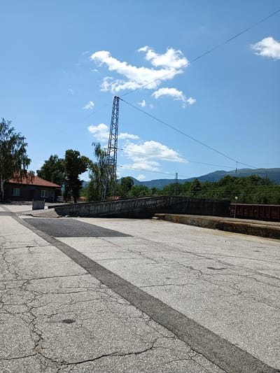 The image captures a view of a section of the Zlatitsa train station in Bulgaria on a sunny day. In the foreground, a cracked asphalt surface is visible, marked with a dark gray stripe. A concrete embankment with graffiti runs diagonally across the midground, sloping downwards from left to right. Beyond the embankment, railway tracks and a maroon freight car are present. In the background, a cluster of trees and buildings, including a small orange-roofed structure with a sign that reads "ЗЛАТИЦА" (Zlatitsa), are situated. Further back, rolling green hills and mountains form the horizon under a bright blue sky with scattered white clouds. A tall, lattice-like metal pylon and overhead power lines are prominent features in the scene. No people are visible in the image.