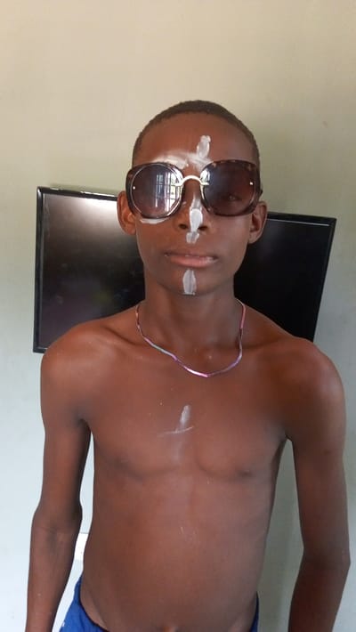 A shirtless, slender, dark-skinned boy wears oversized brown sunglasses. White paint streaks adorn his forehead, nose, mouth, and chest. A thin silver necklace circles his neck. A dark television is visible in the background against a light-colored wall. The boy is facing the camera with a neutral expression. Blue fabric is visible at the bottom of the image.