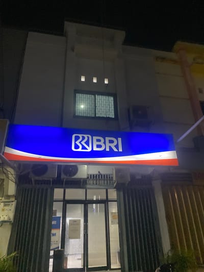 The image shows a nighttime scene in front of a BRI bank branch. A prominent blue and white illuminated sign with the BRI logo is the main subject, with "BRI" clearly visible in white letters. The sign also features red and white stripes on its right side. To the left of the sign, a white building with a window featuring a brick pattern and two square lights is visible. Adjacent to the building, an air conditioning unit is mounted.

To the right of the BRI sign, a dark, slatted shutter covers a storefront. Through the window above the shutter, a second BRI sign with "BRI" and "ATM" visible in blue and orange lettering is partially obscured. Inside the bank, a patterned wall is visible, along with a sign that is too blurry to read. Two air conditioning units are also visible on the exterior wall of the building.

The scene is dimly lit, typical of nighttime. The presence of the BRI bank and the visible ATM sign suggest an urban or commercial setting. There are no people visible in the image. The overall impression is one of a functional, operational bank branch, perhaps in a quiet period at night. The orientation of the image is rotated 90 degrees counter-clockwise.