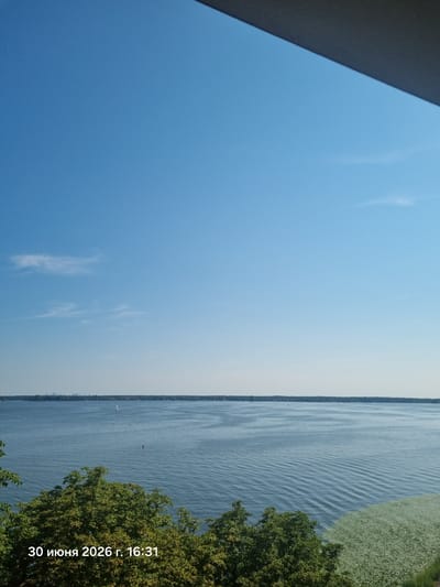 The image shows a wide expanse of a calm, blue lake under a clear blue sky. A thin line of trees and distant buildings marks the far shore. In the foreground, lush green trees partially obscure the lower portion of the scene. A timestamp in the bottom left corner reads "30 июня 2026 г. 16:31", which translates to "June 30, 2026, 16:31" in Russian. This suggests the image was taken in Zegrze, Poland, given the Russian text likely indicates a localized timestamp. The water surface is disturbed by gentle ripples, and a small sailboat is visible in the distance. There are no people or animals clearly identifiable in the image. The overall impression is one of a tranquil, sunny afternoon by the water.