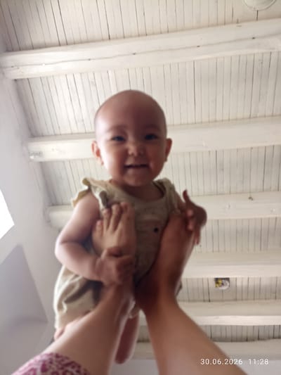 An overexposed, low-angle, wide shot shows a baby being held up by its feet against a white, slatted ceiling. The baby, facing forward, is smiling and blurry, wearing a light-colored outfit. The hands holding the baby are visible at the bottom of the frame. The setting appears to be indoors. A date and time stamp reads "30.06.2026 11:28". The location of Batken City, Kyrgyzstan is not discernible from the image.