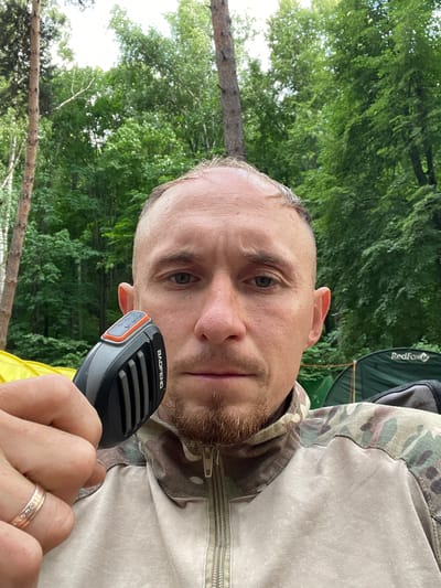 This media file is a close-up, vertically oriented photograph of a man holding a handheld radio in his right hand. The man is positioned in the foreground, and his face is partially visible, turned towards the camera. He has short, light-colored hair with some grey streaks and is wearing a camouflage shirt. A ring with small stones is visible on his right ring finger.
The radio in his hand is black and grey, with the brand name "BAOFENG" clearly printed on it. He appears to be speaking into or listening to the radio.
The background suggests an outdoor setting with lush green trees and foliage, indicative of a forest or wooded area. A portion of a green tent with the "RedFox" logo is visible behind the man's head, and a yellow tarp or tent is also present in the lower part of the frame. The lighting suggests it is daytime, with natural light filtering through the trees.
The overall impression is that of a person engaged in an outdoor activity, possibly camping or hiking, where communication via radio is being utilized. The man's facial expression is neutral, and there are no strong emotional cues. The location context is identified as Tyapino, Russia.