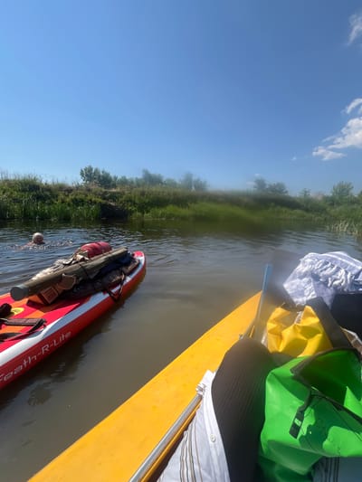 The image depicts a serene river scene on a clear, sunny day. In the foreground, a yellow paddleboard is positioned, laden with dry bags, a towel, and a paddle. To the left, a red and white paddleboard floats in the water, carrying fishing rods and other gear. A person is visible in the water to the left of the red paddleboard, appearing to be swimming or cooling off. The riverbanks are lined with lush green vegetation and trees. The sky is a vibrant blue with a few wispy clouds. The location is identified as the city of Staraya Chigla, Russia.