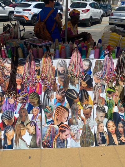 The image captures a vibrant outdoor market stall in Conil de la Frontera, Spain, specializing in hair braiding and accessories. The foreground is dominated by a display of colorful braided hair extensions adorned with beads and feathers, arranged in neat rows. Behind these, large photographic prints showcase diverse individuals with intricate braided hairstyles, ranging from elaborate cornrows to long, flowing braids with vibrant accents.
In the mid-ground, a vendor, a woman with dark skin and a vibrant tie-dye shirt, is actively braiding a young person's hair. She is positioned behind a table laden with spools of colorful thread and other craft supplies, suggesting she is either offering her services directly or is part of the stall's operation. The overall atmosphere is lively, indicated by the abundance of bright colors and the display of various hair artistry.
The background reveals a glimpse of the street environment, with cars parked in the distance and a hint of a building facade, suggesting a typical urban setting. The sunlight is strong, casting shadows and highlighting the textures of the displayed items, contributing to the summery feel of the scene. The stall itself is likely temporary, characteristic of market setups. The arrangement of the photographic prints creates a collage effect, serving as a visual catalog of the available braiding styles and expertise.