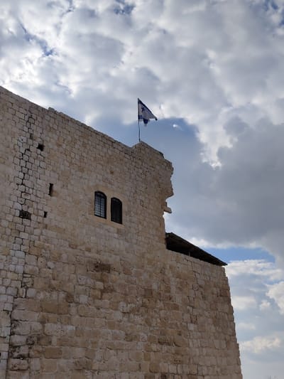 The image displays an upward view of a large, light-colored stone wall, part of the Antipatris Fortress (Qal'at Ras al-Ain) located in Rosh HaAyin, Israel. The wall is constructed from numerous cut stone blocks, exhibiting signs of age and wear. Approximately midway up the visible structure, two narrow, arched windows with dark frames or grates are set into the stone. At the highest point of the visible ruin, an Israeli flag, featuring blue stripes and a blue Star of David on a white field, is mounted on a pole and appears to be gently waving against a backdrop of heavily clouded sky. The clouds are predominantly white and grey, with some patches of blue sky visible. No individuals, specific actions, or interactions are present in the scene.