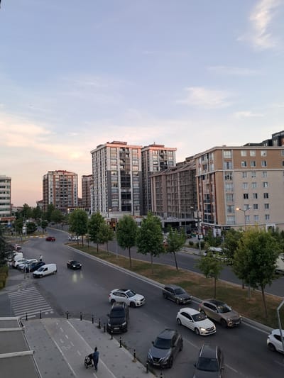 This media file captures a street scene in Beylikdüzü, Turkey, likely during the late afternoon or early evening as suggested by the soft, warm light and the pastel hues in the sky. The image is oriented vertically.
The scene is dominated by several modern, multi-story buildings lining a street. These buildings feature a variety of architectural styles, with some having lighter colored facades and numerous windows, while others appear to be darker or more industrial in design, with some under construction or renovation indicated by scaffolding. Trees with lush green foliage are interspersed between the buildings and along the roadside, contributing to a sense of urban greenery.
The street itself is paved and appears to be a main thoroughfare. Several cars are parked along the street and in motion. Visible vehicles include a white sedan, a dark-colored SUV, and other various sedans and hatchbacks. One person is visible on the sidewalk, pushing a trolley.
The sky is a pale blue with wisps of white clouds, hinting at fair weather. The overall atmosphere is calm and urban, with a focus on the interplay between the modern architecture, the natural elements of trees, and the everyday activity of a city street. There is some visible text on one of the buildings, appearing to be "ARIFOGLU".