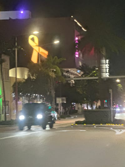 This night-time image depicts an urban street scene in Miami Beach, United States, specifically identifiable by the prominent illuminated yellow ribbon-shaped sign characteristic of Lincoln Road.
In the upper center-left, a large, yellow, illuminated ribbon-shaped sign is suspended, marking a prominent feature of the environment. Directly behind and to the right of this sign, a building features a vertical pink neon sign spelling "PALACE" and another vertical sign with white neon letters, partially visible as "SSIS" or similar. Streetlights illuminate the area, some creating lens flares. Lush green foliage, possibly palm trees, is visible.
In the middle ground, a dark-colored SUV or truck is captured in motion on the roadway, facing away from the viewer, with its bright headlights illuminated. The vehicle exhibits slight motion blur. Further down the street on the right, additional faint vehicle lights are discernible. The roadway in the foreground features white lane markings and a directional arrow. A central median or planter divides the road, adorned with multiple yellow road markers (cat's eyes). Blurry figures are present on the sidewalk in the background, indicating pedestrian activity. The overall scene suggests a vibrant commercial district at night.