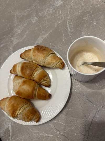 Four croissants are arranged on a white plate. To the right of the plate, a white mug contains a frothy beverage, likely coffee or cappuccino, with a spoon in it. The subjects are placed on a gray marble-patterned surface. The image does not provide any information about the location in Murom, Russia.