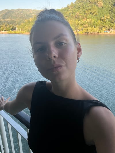 A close-up, eye-level selfie shows a woman with her hair pulled back in a bun, looking directly at the camera. She is wearing a black sleeveless top and a silver hoop earring in her right ear. Her left hand is resting on a white railing. In the background, there is a body of water and a lush green hillside with palm trees and some buildings. The scene appears to be set in Batas Desa Skotong Timur, Indonesia, likely on a ferry or boat as indicated by the railing and the open water. The sun is shining, casting a warm light on the scene.