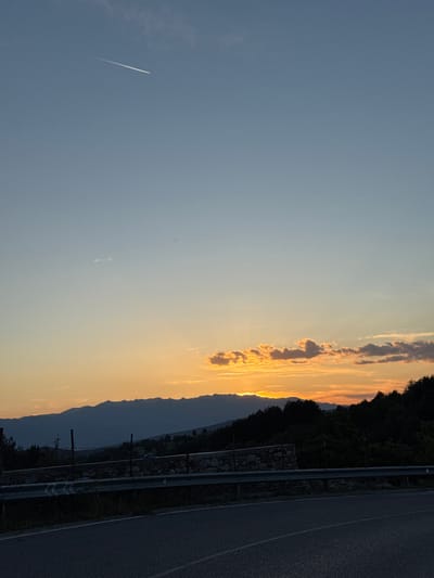 A sunset over a mountainous landscape near Valkosel, Bulgaria, is captured in this image. The sky transitions from a clear blue at the top to shades of orange and yellow near the horizon, illuminated by the setting sun. Wispy clouds are present, and a faint contrail from an airplane is visible in the upper portion of the sky. Silhouetted against the vibrant sky are a range of mountains, their peaks softly defined. In the foreground, a dark line of trees and foliage can be seen, with a low stone wall and fence running along the edge of a paved road. The road curves gently, its surface dark and textured. The overall scene depicts a serene natural landscape during twilight.