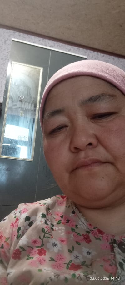 A close-up, slightly tilted selfie shows a middle-aged woman wearing a pink head covering and a light-colored blouse with a floral pattern featuring pink, white, and red flowers. She has dark eyes and a somewhat rounded face with visible wrinkles around her eyes and mouth. Her expression appears to be a slight grimace or squint. To the left of the woman's face, a full-length mirror is partially visible, reflecting what appears to be an indoor setting with some objects and lights within the reflection. The background wall has a textured pattern. A date stamp "23.06.2026 14:48" is present in the bottom right corner of the image, indicating the time and date the photo was taken. The location of Chon-Kara, Kyrgyzstan is not depicted in the image, and no elements within the visual content confirm this location.