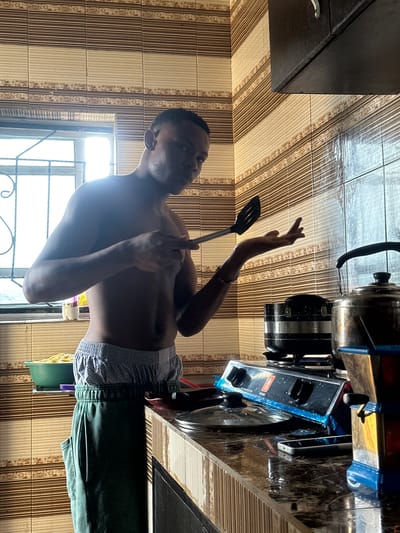 A shirtless man stands in a kitchen in Obinze, Nigeria, holding a spatula. He wears patterned boxer shorts and green sweatpants. Behind him is a window, and to his right is a stove top with pots on it. The kitchen has brown-striped tile walls.
