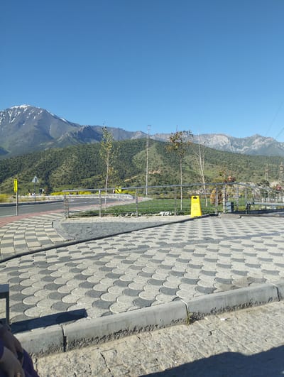 The image shows a paved area with a patterned walkway in the foreground, leading towards a road. Beyond the road, a metal railing separates the paved area from a grassy field dotted with yellow flowers. In the background, a lush green hillside rises, culminating in a majestic mountain range with snow-capped peaks under a clear blue sky. The scene appears to be a scenic overlook or park area in Dardar, Uzbekistan, given the presence of mountains and possibly local flora. There are several trees, including one with light-colored leaves, scattered across the landscape. A yellow cautionary sign is visible near the railing, and a few individuals can be seen walking near the road in the middle ground. The overall impression is one of a serene natural environment with man-made pathways and infrastructure.
