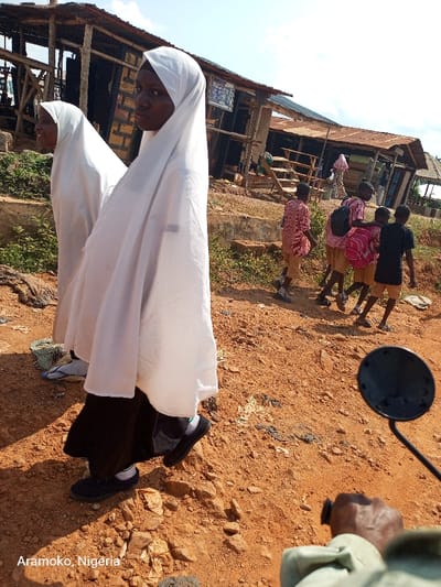 The image shows a street scene in Aramoko, Nigeria, located at latitude 7.836 and longitude 4.476. Two young girls are walking in the foreground, both wearing white hijab, one of them wearing black trousers and the other wearing black and white trousers. There are five children walking in the background, all wearing school uniforms and shoes. They are walking away from the viewer, on a dirt road. There is a small building in the background, with a wooden structure in front. The ground is dirt and there is some vegetation on the side of the road. The image is taken from a low angle, facing the girls walking. The viewer's hand is visible in the foreground holding the handle of a motorbike. The sun is shining brightly in the background.