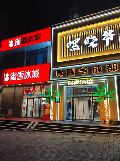 This image captures the exterior of two businesses at night, located in Jiguan District, China. The building features brightly lit signage. On the left, a red sign with white Chinese characters, "蜜雪冰城" (Mi Xue Bing Cheng), is prominently displayed, indicating a franchise known for ice cream and drinks. Below this sign, the store entrance is visible, with a large cartoon character in front of it. To the right, a larger, yellow-gold illuminated sign with white Chinese characters, "嘿晓爷" (Hei Xiao Ye), and "深夜酒馆" (Shen Ye Jiu Guan - Late Night Bar) is displayed. This establishment has a glass entrance flanked by decorative palm trees made of lights. The pavement in front of the stores is paved with cobblestones. The overall scene suggests a commercial street illuminated at night.