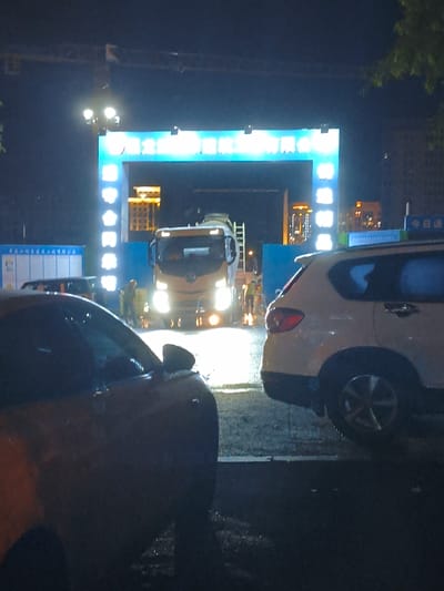 In the city of Jiguan District, China, a construction site is visible at night. The scene is illuminated by a large, blue archway with bright white text that reads "Jiguan District". Two cars are parked in the foreground, one red and one white SUV. In the background, a cement truck is positioned under the archway. A construction worker in a yellow vest and hard hat is standing near the truck, holding a shovel. A tower crane is also visible in the distance, suggesting ongoing construction work. The ground is wet, indicating recent rainfall. The overall atmosphere is dark and industrial, typical of a construction site at night.