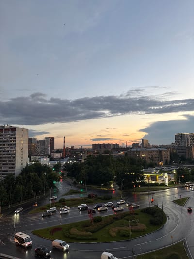 This is a high-angle, slightly rotated, wide shot of a city intersection during dusk. The sky is a mix of light grey clouds and a warm orange and yellow sunset. Tall buildings, some with illuminated windows, line the sides of the road. Trees are also visible, creating a green canopy.
The main focus is a multi-lane road with several cars driving on it. The road appears wet, suggesting it has recently rained or is still raining lightly. Traffic lights are visible, and some are red, indicating traffic is stopped. A circular traffic island with manicured bushes and trees is at the center of the intersection. There is a prominent red star-shaped structure on the traffic island.
In the distance, a tall brick chimney with a red cap stands out against the skyline. The overall mood is calm and serene, with the soft lighting of dusk and the quiet hum of the city. The location is identified as Chelyuskinsky, Russia.
