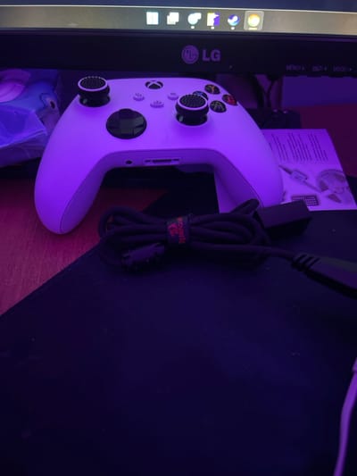 A white Xbox controller with black thumbsticks is resting near a monitor. A black Redragon-branded cable is coiled next to it. The objects are illuminated by purple lighting. Part of an LG monitor and a paper sheet are visible behind the controller. The scene is set on a dark-colored mousepad.