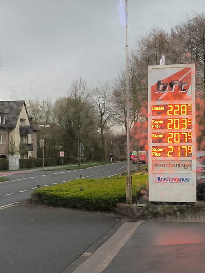 The image depicts a street scene in Havixbeck, Germany, on an overcast day. A BFT gas station sign dominates the right side of the frame, displaying prices for Diesel at 2.289, Super E10 at 2.039, Super at 2.079, and Super Plus at 2.179. "Waschanlage" (car wash) and "Autogas" are also displayed. A white flag on a pole stands behind the sign.
A two-story building is visible on the left side of the frame, across a two-lane road. A green hedge borders the road and a median is covered in green foliage. A person is walking on the far side of the street. A red car is pulling into the gas station. The image appears to have water droplets on the lens.