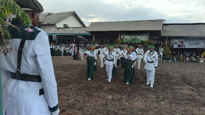 The image shows a group of people in white and green uniforms standing in formation in an open area in front of a building in the city of Keshero, Democratic Republic of the Congo. The building has a corrugated metal roof and appears to be partially constructed. There are other people watching from the sidelines. One person in the foreground is wearing a green beret and white and green uniform, standing facing to the left of the frame. The people in the formation are performing a drill or marching routine. The ground is dirt and there is foliage in the foreground. There is a banner hanging on the building that says "G.T.K." and "KOKO KIVU". There is also a poster with a picture of a man on it. The photo appears to have been taken during a formal event, perhaps a ceremony or parade. The mood is one of order and formality. The event appears to be taking place in a public space, perhaps a schoolyard or a community gathering area. The event appears to be related to the "G.T.K.", which could be a school, organization, or community group.