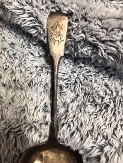 A close-up overhead shot shows a tarnished silver spoon resting on a grey, fluffy fabric. The spoon has a monogram etched into its handle. The fabric appears to be a blanket or rug, with long, wispy fibers. The background is out of focus, emphasizing the spoon and fabric. No people or specific location details within the city of Ново-Талицы, Russia are visible.