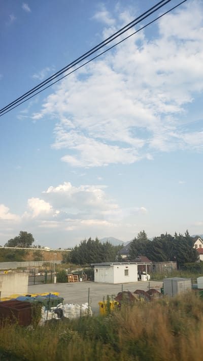 The image depicts an outdoor scene on a bright, cloudy day, likely in Bešeňová, Slovakia. In the foreground, overgrown grass and vegetation are visible. Behind this, a fenced-off area contains various objects, including several large industrial bins of different colors (brown, blue, green, yellow), stacks of white bags, wooden pallets, and some rusty metal debris.
Further into the scene, a small, white modular building with a window and a door stands on a paved surface. Adjacent to it is a smaller, dark-roofed structure. A few people can be seen near the white building, and a washing machine is positioned outside of it. Behind these structures, a dense line of dark green coniferous trees forms a backdrop. Beyond the trees, a few houses with dark roofs are visible, and in the far distance, mountains with patches of snow can be seen under a vast blue sky filled with scattered white clouds. Overhead, three dark power lines stretch across the upper portion of the image. The overall impression is that of an industrial or storage area bordering a residential zone, set against a natural landscape.