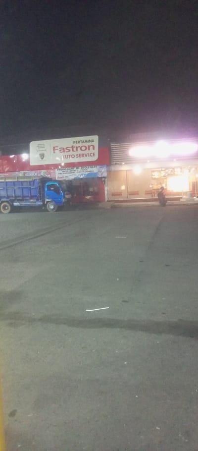 A blue truck is parked in front of a Pertamina Fastron Auto Service center at night. The auto service center advertises a "PROMO GANTI OLI DI OLI MART" (oil change promotion) with "cheer's" branding. To the right of the auto service center is a donut shop with illuminated displays. A person on a motorcycle is visible near the donut shop's entrance. The ground is paved and dimly lit. The night sky is dark.